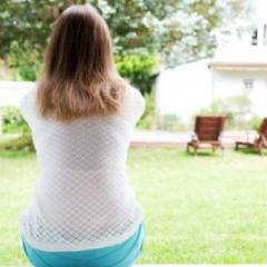 A woman sits at an open doorway with her back to the camera. She is facing a garden with a green lawn, trees and wooden chairs.