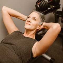 An older woman in her 50's, working out at the gym on equipment, looking up with a smile.