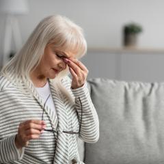Woman sitting on a grey couch holding her glasses and touching her temple.