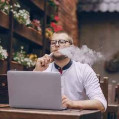 Man smoking vape while sitting at table with laptop open in front of him 